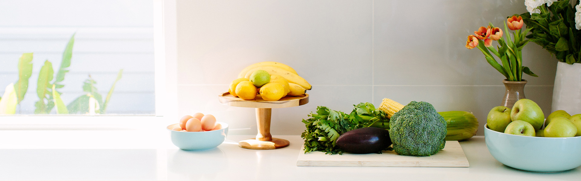 Fresh fruit and vegetables with a bowl of eggs and vase of flowers on a kitchen bench